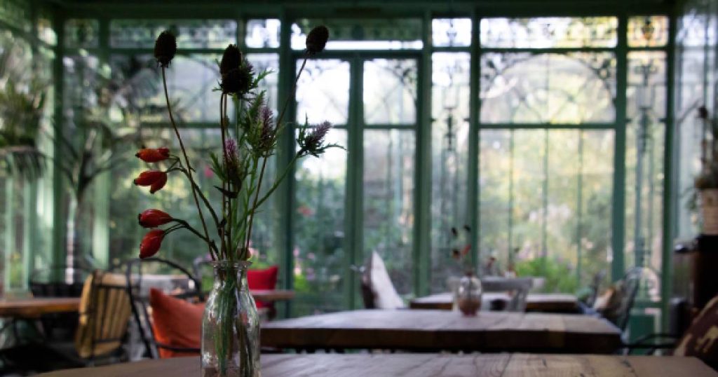 Het interieur van een smeedijzeren serre met grote raampartijen en smalle, elegante profielen. Op een tafel in de serre is een glazen vaas met wat bloemen erin te zien.