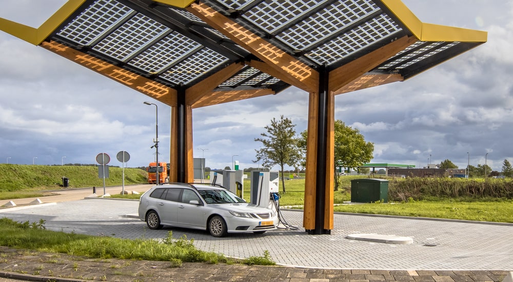 Een elektrische auto aan een laadpaal naast een snelweg in een groen gebied. De auto staat onder een grote, abstracte gele pergola.