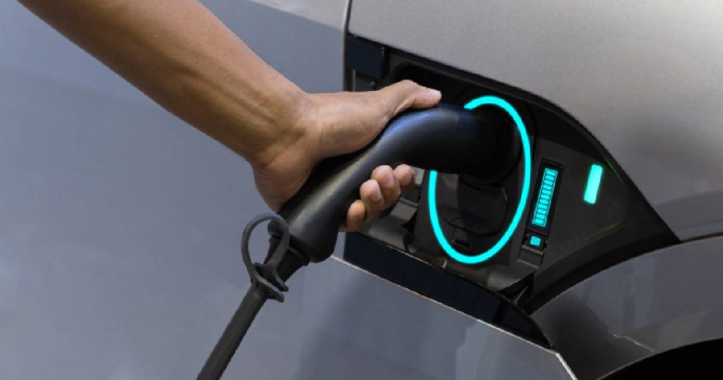 Detailbeeld van een hand die een laadkabel van een elektrische wagen vasthoudt tijdens het opladen. Een cirkel van groen licht rond het aansluitpunt geeft aan dat de koppeling tussen de wagen, laadkapel en een van de verschillende soorten laadpalen geslaagd is.