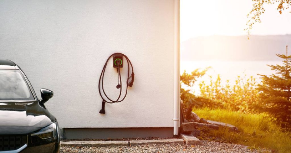 Een laadpaal in vorm van een wallbox, een van de verschillende soorten laadpalen voor thuis, tegen de witte gevel van een privéwoning. De vaste laadkabel hangt rond de box, naast de laadpaal staat een zwarte elektrische wagen. Op de achtergrond een zonnig landschap met planten, meer en berg.