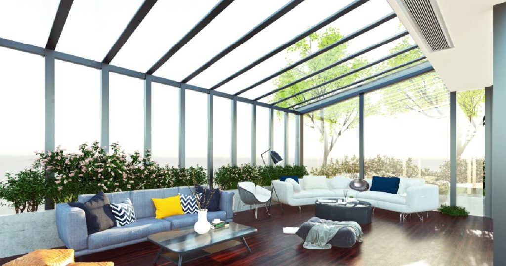 Een moderne stalen serre met lange, smalle profielen en een helder uitzicht op de omgeving. In de serre ligt een houten vloer en staan verschillende zitmeubels.