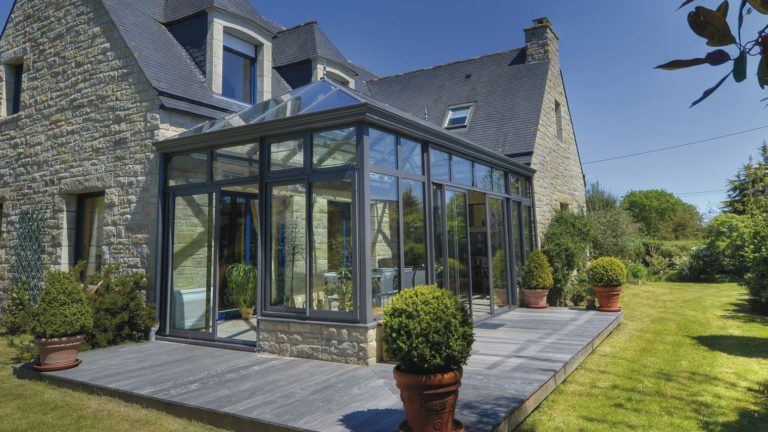 Serre aan huis uitgevoerd met grote raampartijen en schuifdeuren. In de serre staat een tafel met stoelen en planten.