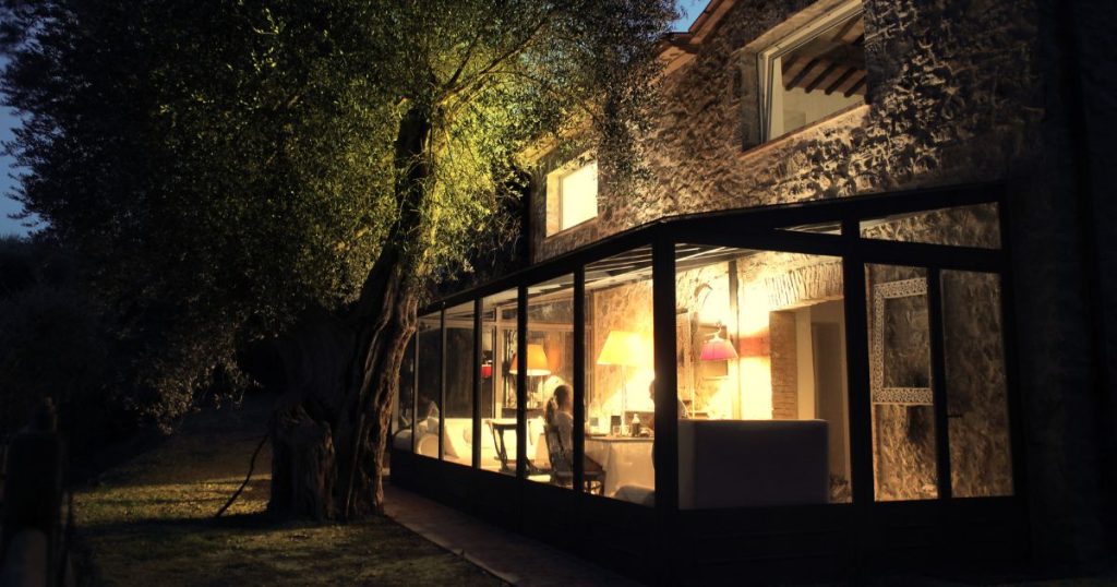 Grotere glazen serre waar wel vergunning voor nodig is. De serre grenst aan een stenen huis en in de verlichte serre is een persoon te zien met meubilair