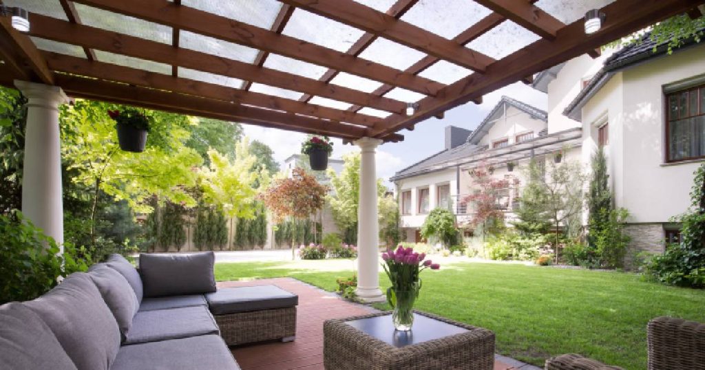 Een vrijstaande pergola met witte pilaren en een melkwit polycarbonaat dak in houten frame. Onder de open veranda staat een loungeset met grijze kussens en een bijzettafeltje met vaas.