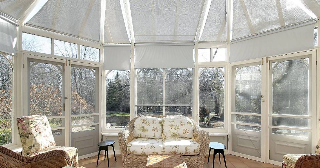 Een serre met lichtkoepel en zonwering. In de serre staat een rieten stoel met witte kussens met bloemmotief, twee zwarte krukjes en een rieten tafeltje.