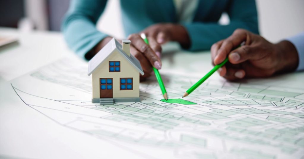 Een bouwplan met groene perceelaanduiding. Op het plan staat een klein modelhuisje. Twee mensen wijzen eenzelfde plot aan met felgroene potloden. Enkel hun handen en armen zijn in beeld.