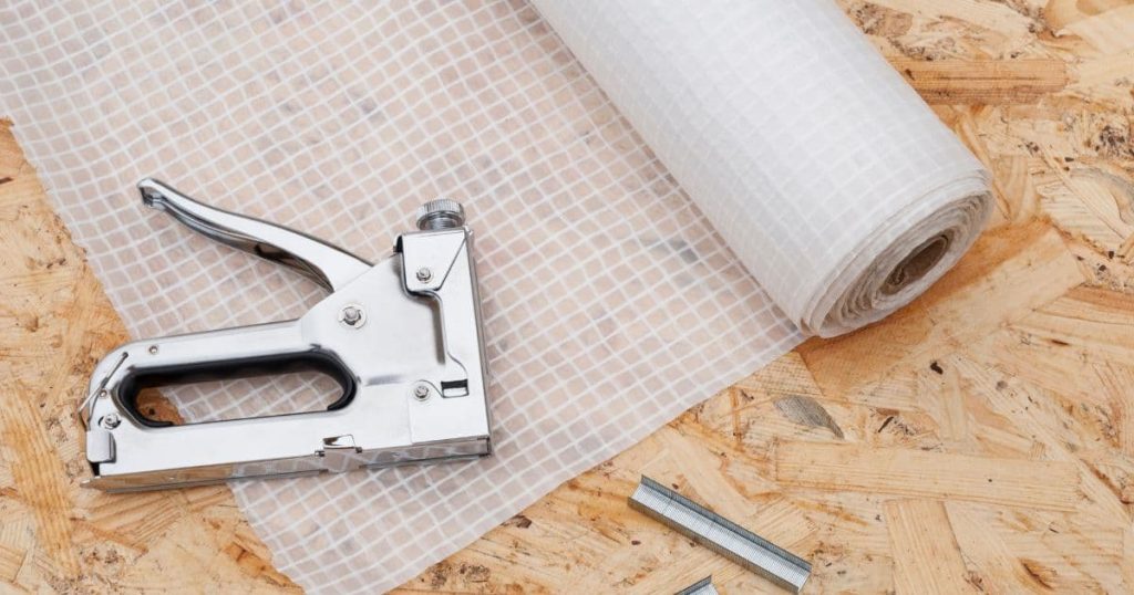 Een rol transparante gevelfolie of geveldoek met wafelstructuur op een houtvezelplaat. Op het afgerolde stuk folie ligt een nietpistool. Naast de folie liggen 2 rijen met nietjes voor het nietpistool.