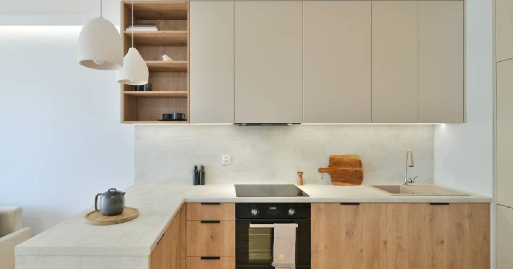 Een keuken in Japandi stijl met gebroken witte bovenkasten, houten onderkasten en een wit werkblad met ingebouwde kraan in een iets donkerder gebroken witte tint. Twee designlampen hangen boven het schiereiland dat verbonden is met het keukenblok onder de bovenkasten.