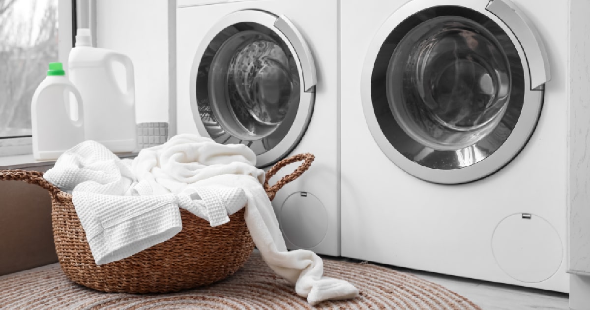 Een rieten wasmand met wit wasgoed staat voor een witte droogkast en witte wasmachine die door zonne-energie uit een thuisbatterij worden aangedreven.