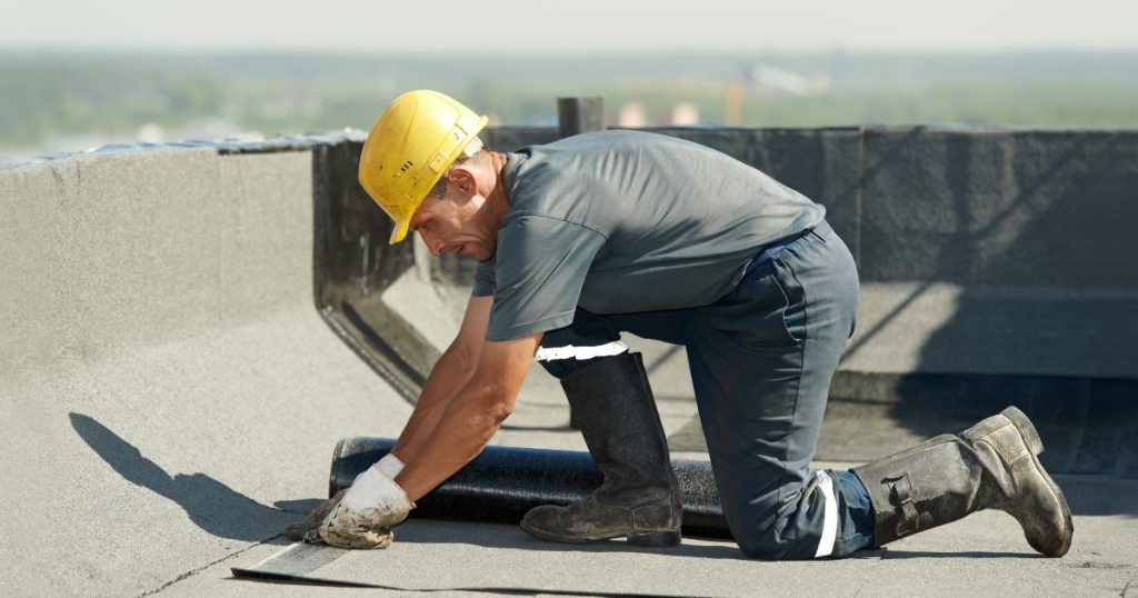 Een isolatiespecialist in grijs T-shirt, werkbroek, laarzen en gele veiligheidshelm werkt een plat dak af dat net geïsoleerd werd met PIR-dakisolatie.