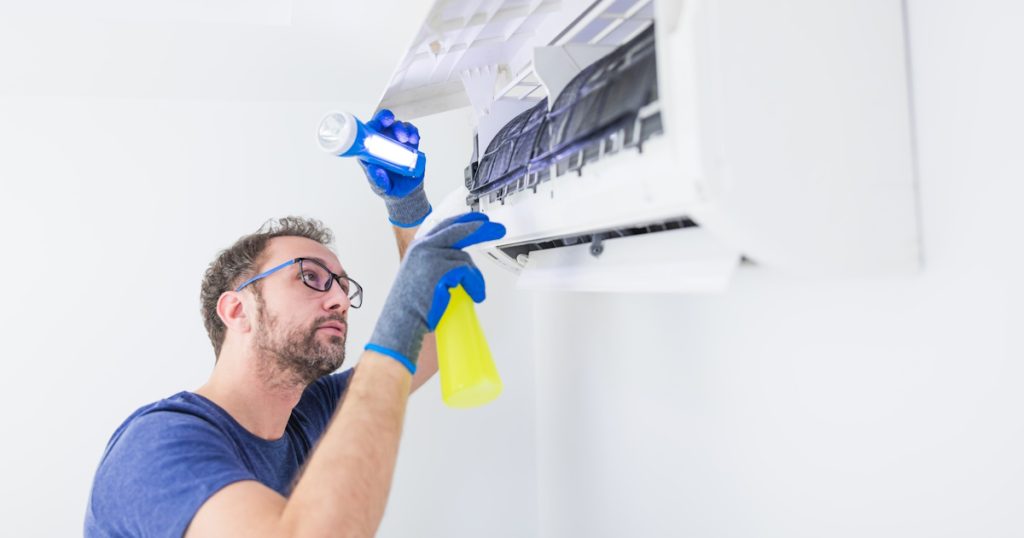 Een man reinigt de binnenunit van een airco. Hij gebruikt hiervoor een zaklamp en draagt beschermende handschoenen.