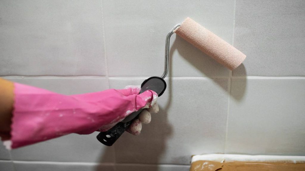Een hand met een roze handschoen schildert de tegels van een badkamerwand met een verfroller. 