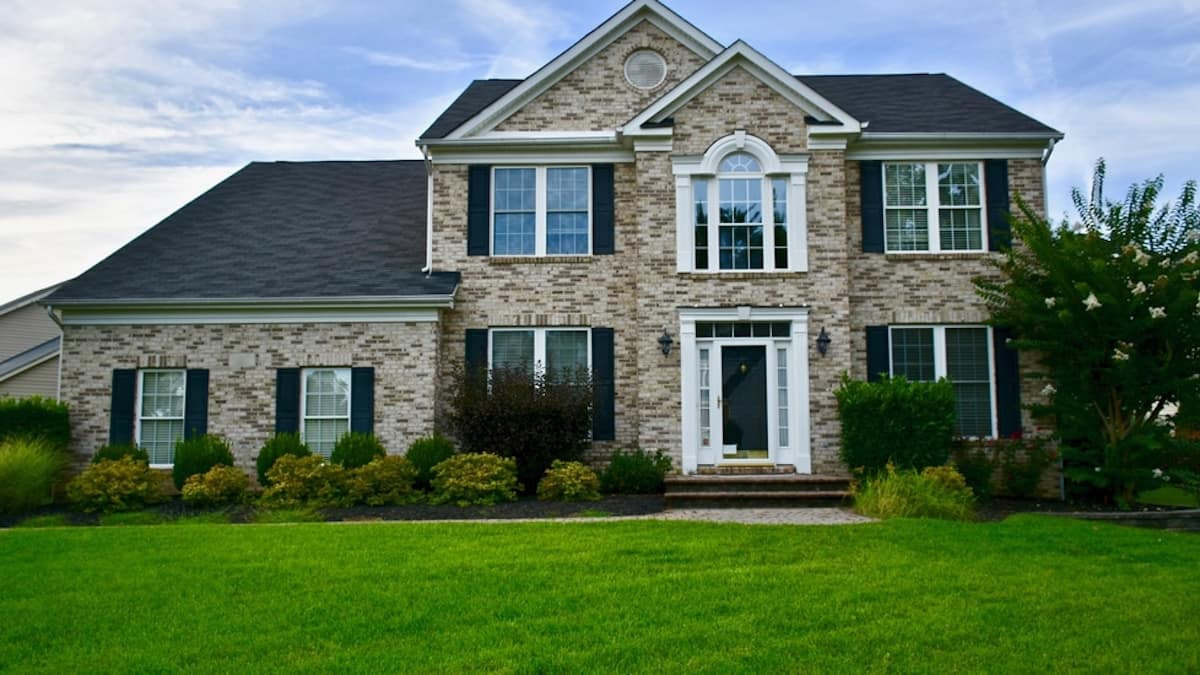 De gevel van een vrijstaande woning. De bakstenen in de gevel variëren van lichtgrijs tot bruin. Op de voorgrond zijn een boom, struiken en een grasveld te zien.