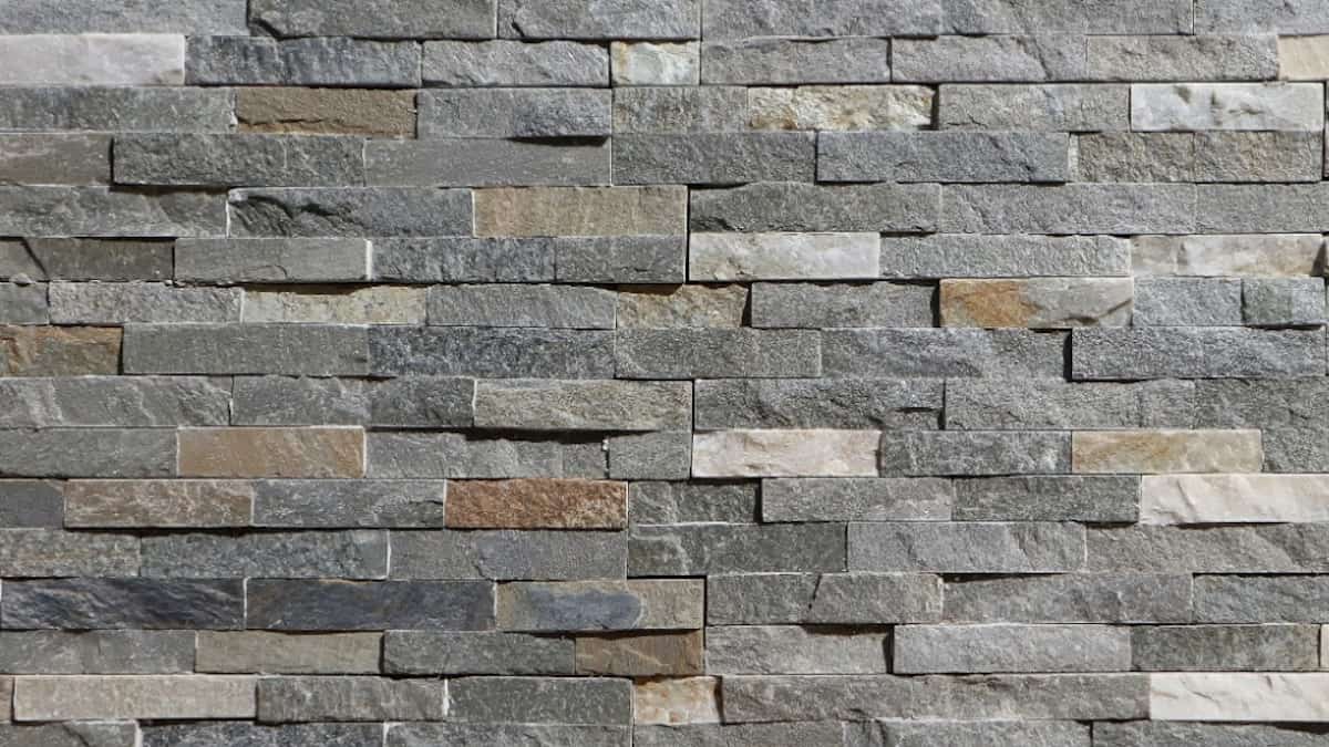 Een gevel van bakstenen. De bakstenen zijn grijs, beige en bruin. De muur is niet egaal, maar juist onregelmatig.