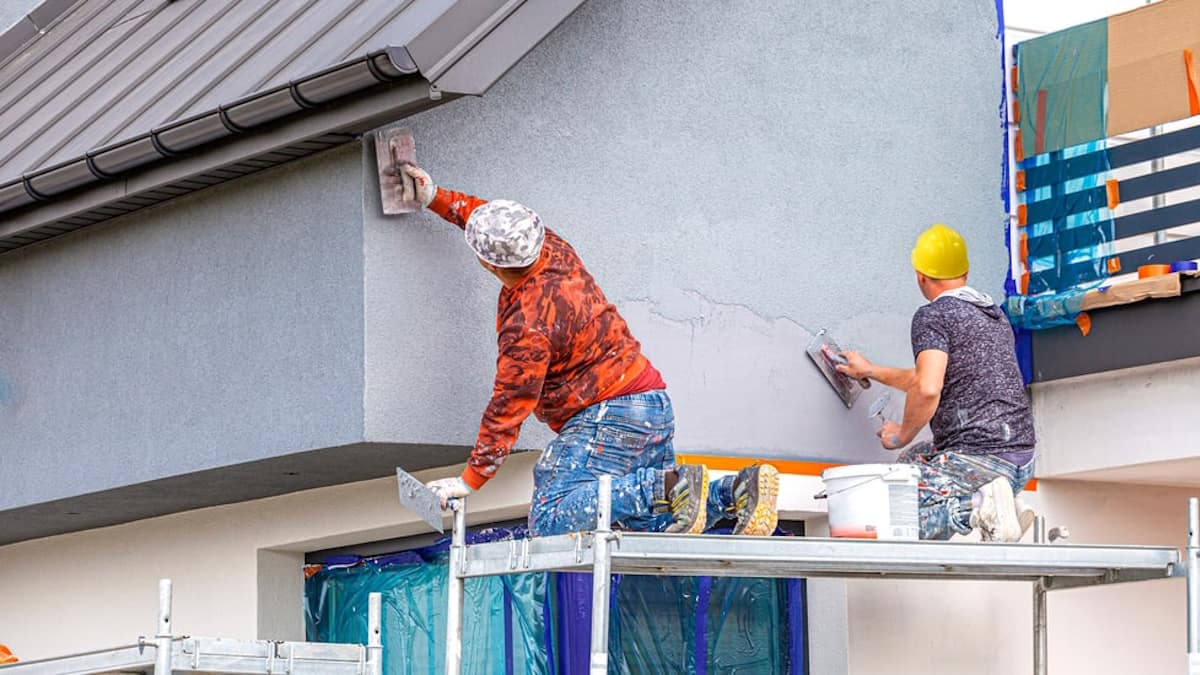 Twee stukadoors brengen sierpleister aan op de gevel van een woning. Ze werken op een steiger en dragen veiligheidshelmen.