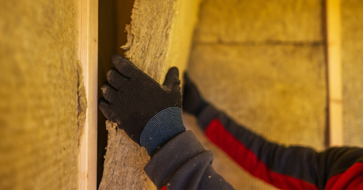 Een isolatiespecialist plaatst een rol minerale wol tussen houten latten. De expert draagt beschermende kleding om irritatie van de vezels te voorkomen