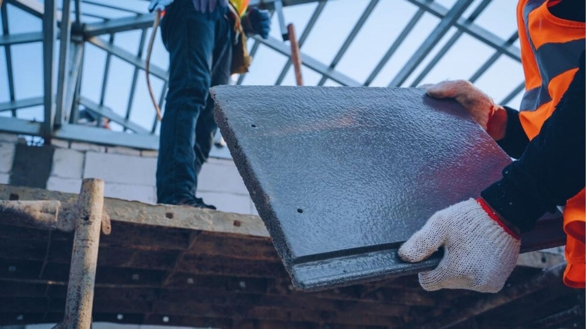 Een dakwerker met fluohesje en werkhandschoenen houdt een betonnen dakpan vast: een soort dakbedekking met een levensduur van 30 tot 50 jaar.