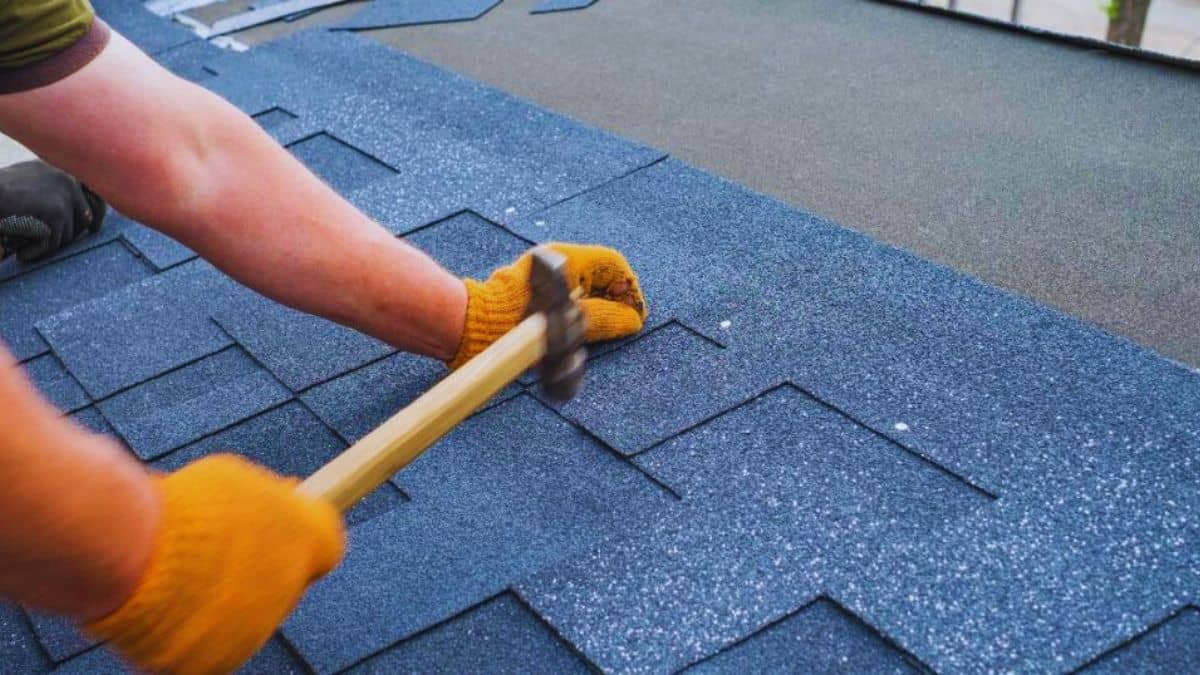 Een dakspecialist gebruikt een hamer en asfaltnagels om bitumen dakshingles op een licht hellend dak te plaatsen.