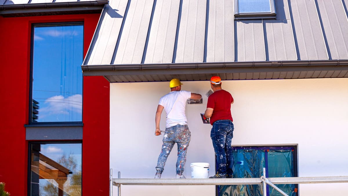 Twee stukadoors zijn bezig met de afwerkingslaag van de sierpleister op de gevel. Het gebouw heeft aan de linkerzijde een opvallend rode kleur.