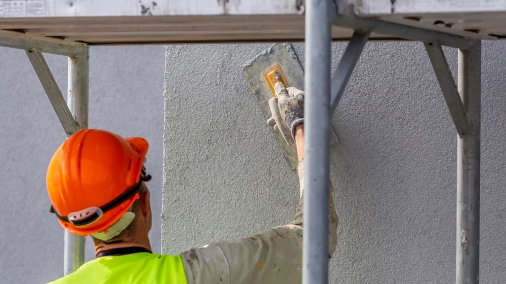 Een stukadoor met beschermende kleding bepleistert de gevel. De man draagt beschermende kleding en staat onder een steiger.