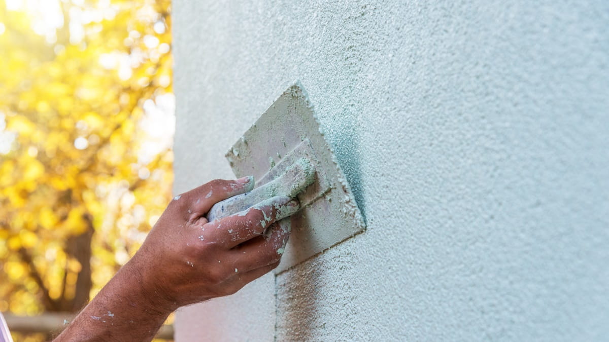 Een stukadoor bepleistert een buitengevel met crepi. De stukadoor gebruikt er een spaan voor. Op de achtergrond is een boom te zien.