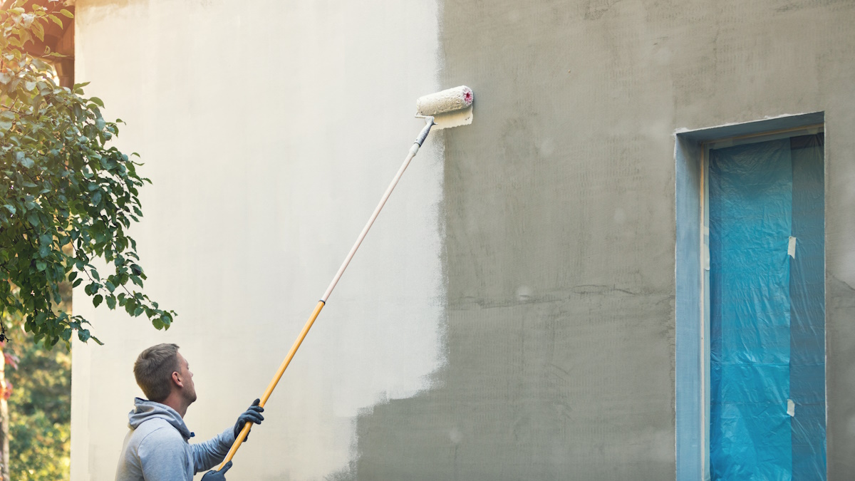 Een schilder verft de gevel van een gebouw. De gevel is grijs en wordt in een lichte kleur geschilderd. De schilder gebruikt een telescoopsteel met verfroller en draagt donkere handschoenen.