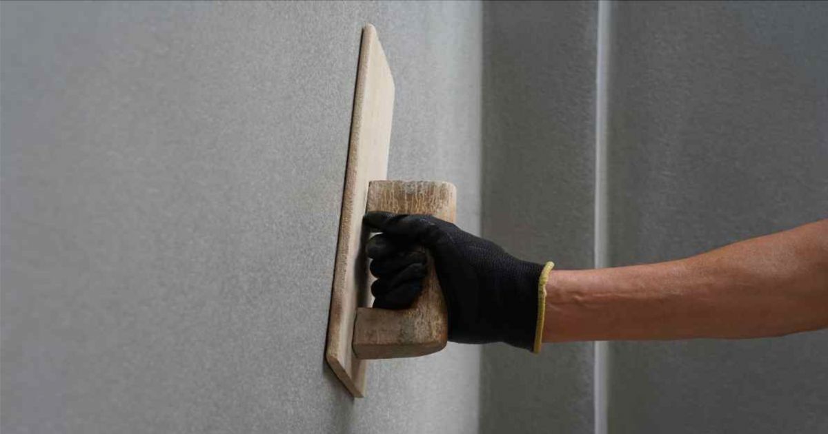 Een vakman met een zwarte handschoen brengt cementpleister egaal aan op een buitenmuur