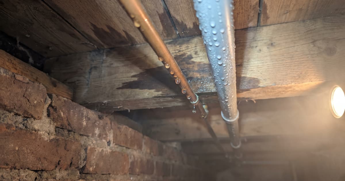 Condensdruppels hangen aan een koperen en een grijze leiding die onder de houten vloerbalken van een kruipruimte lopen, met donkere vochtplekken op het hout en een oude bakstenen muur ernaast.