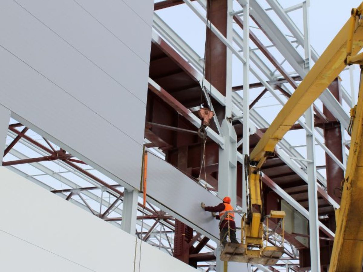 Een constructiewerker installeert sandwichpanelen op een stalen gebouwstructuur met behulp van een kraan en hefplatform.