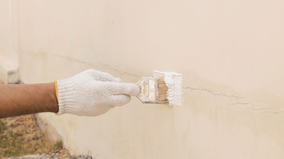 Een schilder verbergt een haarscheur in crepi door de scheur te schilderen met witte verf. De man gebruikt een blokkwast en draagt een witte handschoen.