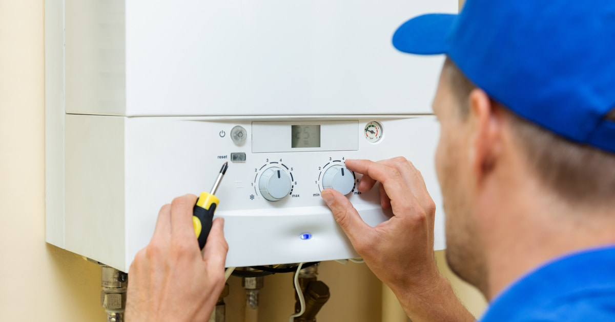 Een monteur draait aan de knoppen van een cv-ketel. De monteur draagt een blauwe pet, een blauw shirt en heeft een schroevendraaier vast