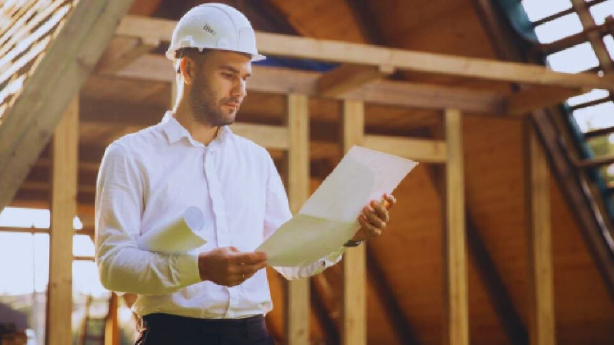 Een architect in wit hemd, witte veiligheidshelm en zwarte broek bekijkt de plannen voor het verhogen van een dak.