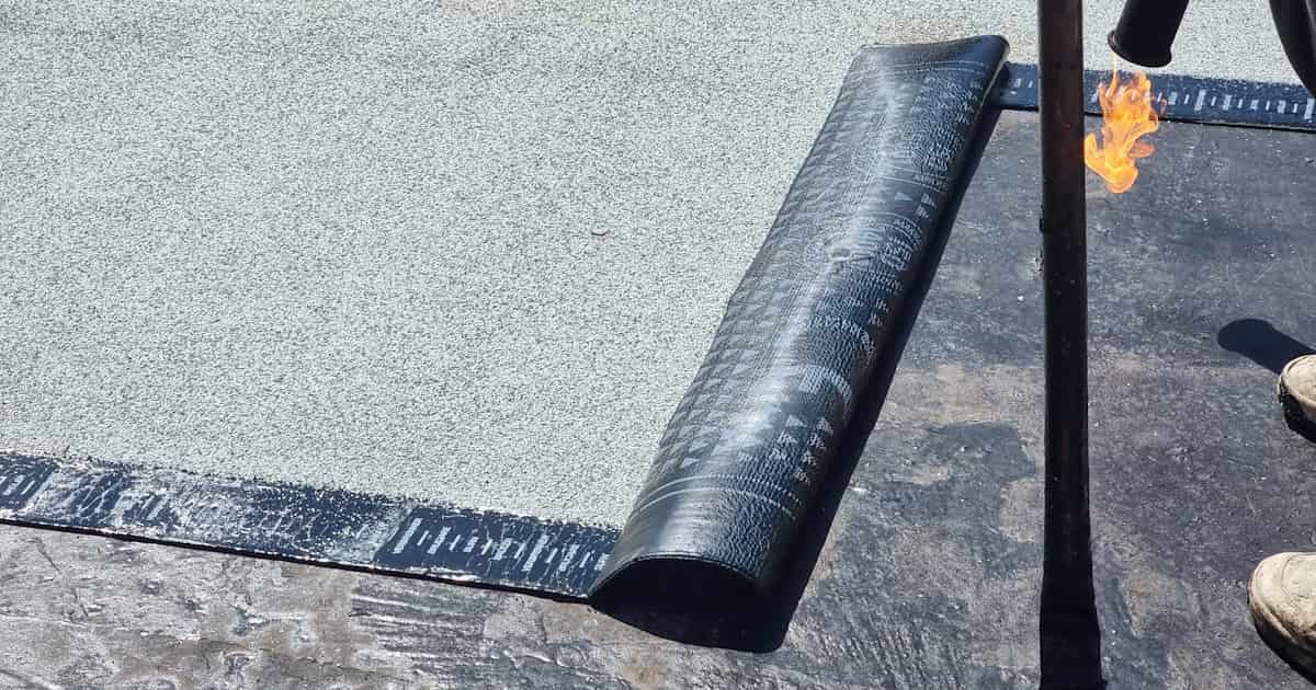 Een dakdekker gebruikt een gasbrander om een strook bitumen dakbedekking op een plat dak aan te brengen