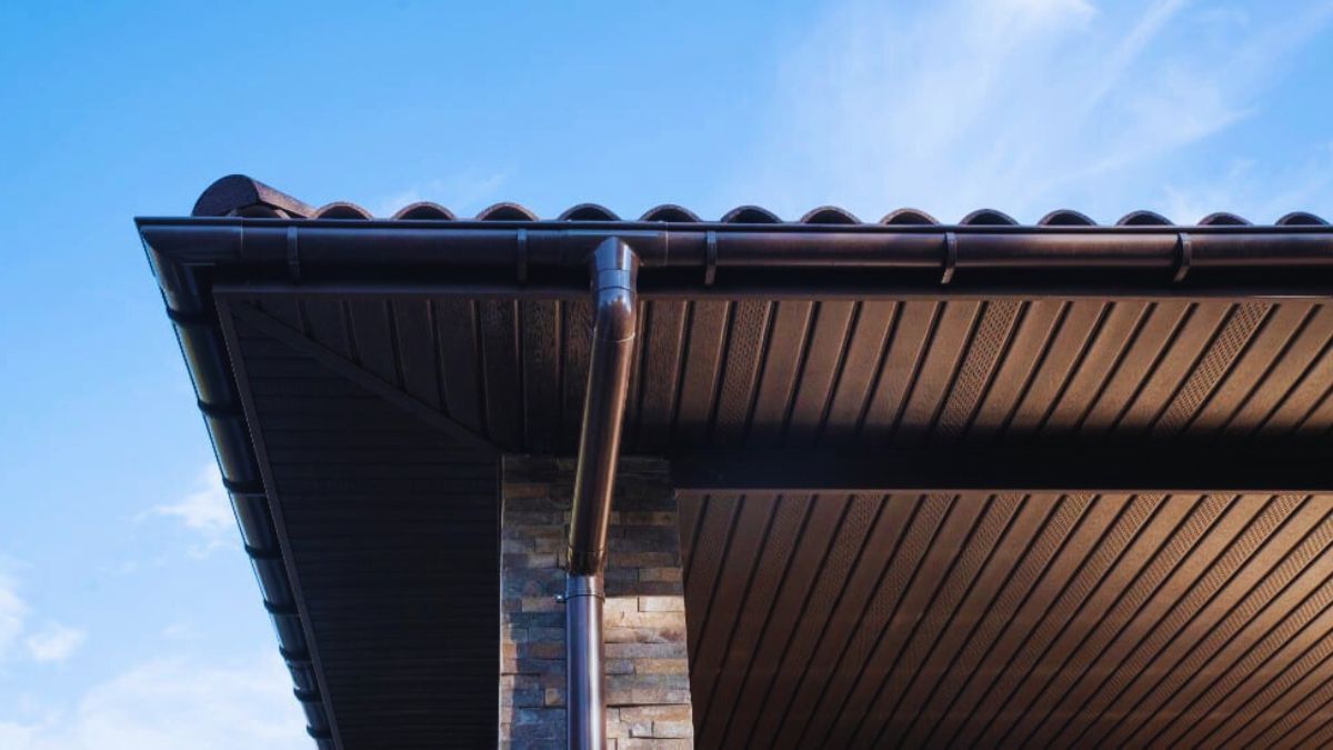 Een dakoverstek aan de onderkant van een dak met dakpannen. Ook is er een donkere dakgoot te zien met een regenpijn eronder.
