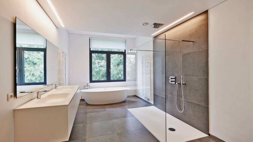 Een ruime badkamer met een wit bad, witte wastafels en een ruime inloopdouche met grijze wandtegels. 