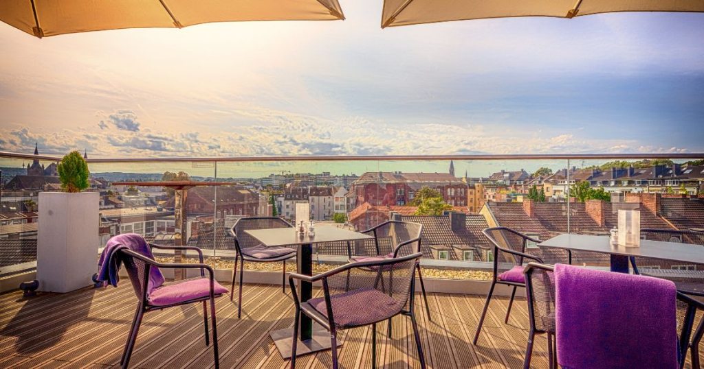 Een dakterras met 6 stoelen en 2 tafels onder twee parasols, op een houten vloer die rust op drukvaste isolatieplaten. Het terras heeft uitzicht op de daken van lager gelegen woningen in een dorp. Op de achtergrond een lichtblauwe hemel met wolken.