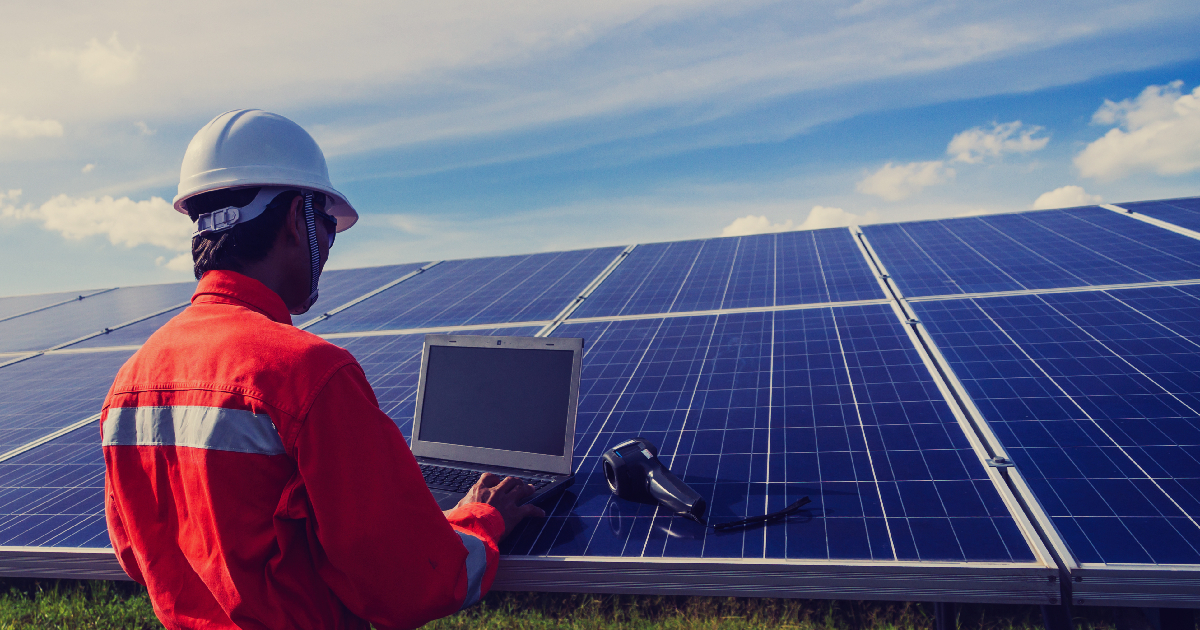 Een ervaren installateur in een rode jas en een witte helm staat, met zijn computer in de hand, een installatie met zonnepanelen te controleren.