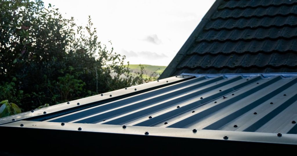 Een dak met dakpannen loopt uit in het dak van een uitbouw dat bedekt werd met geïsoleerde dakplaten. De woning ligt in een groene omgeving.