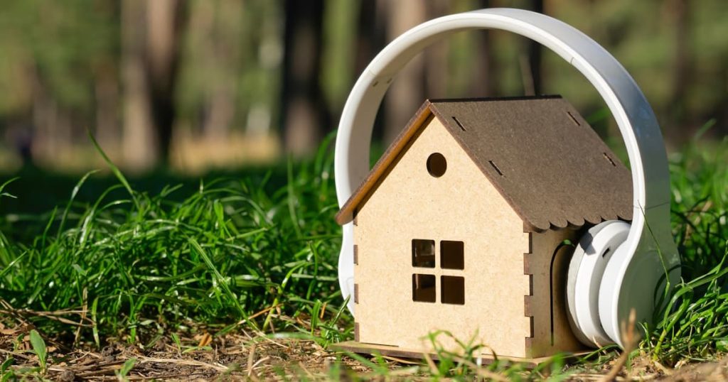 Een houten huisje draagt een hoofdtelefoon tegen geluidsoverlast. Op de achtergrond gras en bomen