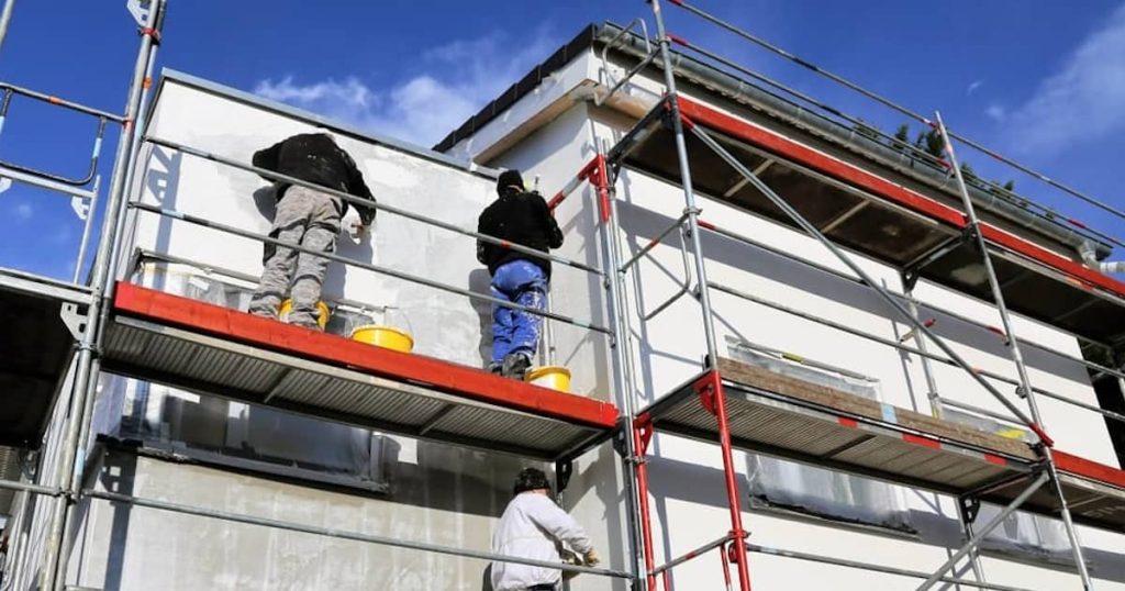 Drie gevelspecialisten schilderen de gevel van een gebouw. Rechts is de nieuwe, frisse gevel zichtbaar terwijl de gevel links nog vervuild is.