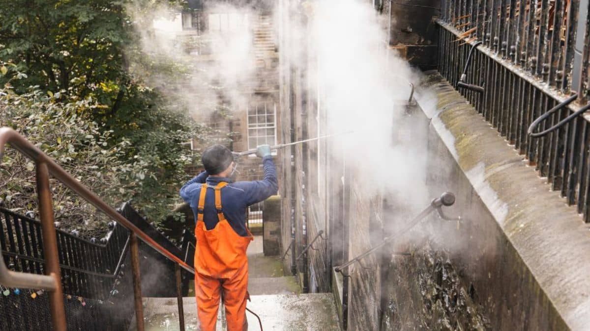 Professionele reiniger stoomt een vervuilde gevel schoon met een stoomreiniger in een steeg met trappen.