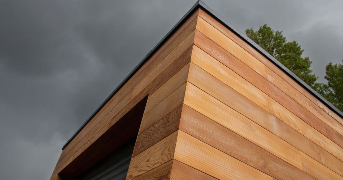 Een gevel met sidings gemaakt van hout. Het hout heeft verschillende tinten bruin. Op de achtergrond een donkere lucht.