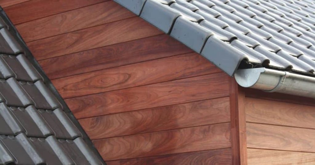 Een gevel gemaakt van houten sidings. Het hout heeft een warme kleur. Op het dak liggen donkere dakpannen.