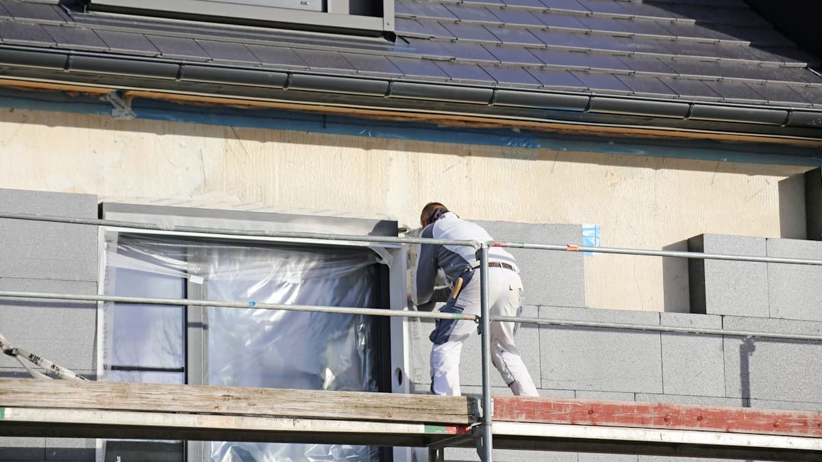 Een gevelwerker isoleert de gevel van een huis. De gevelwerker staat op een steiger en draagt beschermende kleding. Op het dak zijn grijze dakpannen zichtbaar.