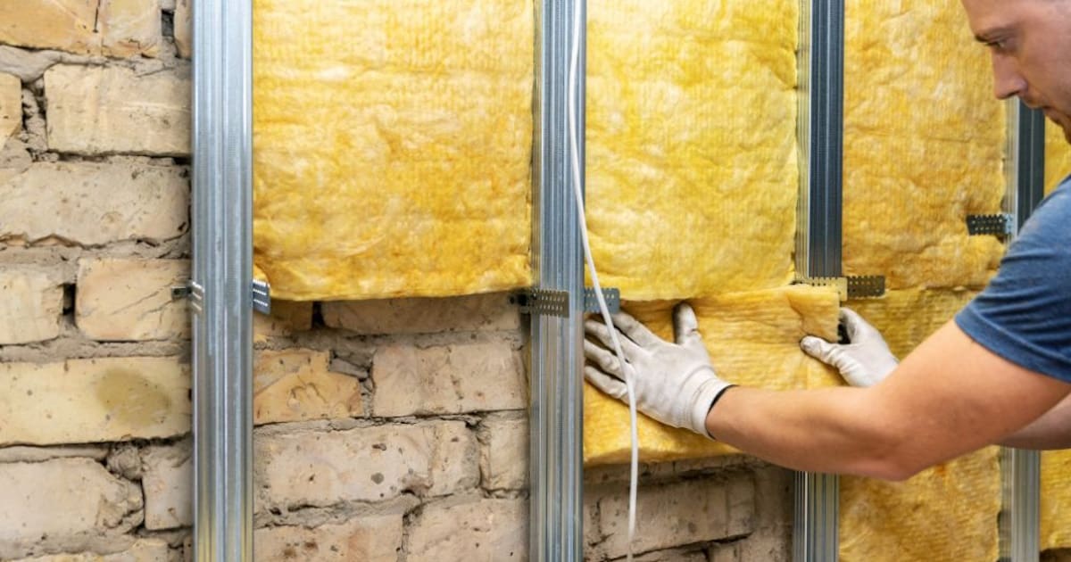 Een isolatiespecialist plaatst minerale wol aan de binnenzijde van een woning. De expert draagt beschermende handschoenen en een blauw shirt.