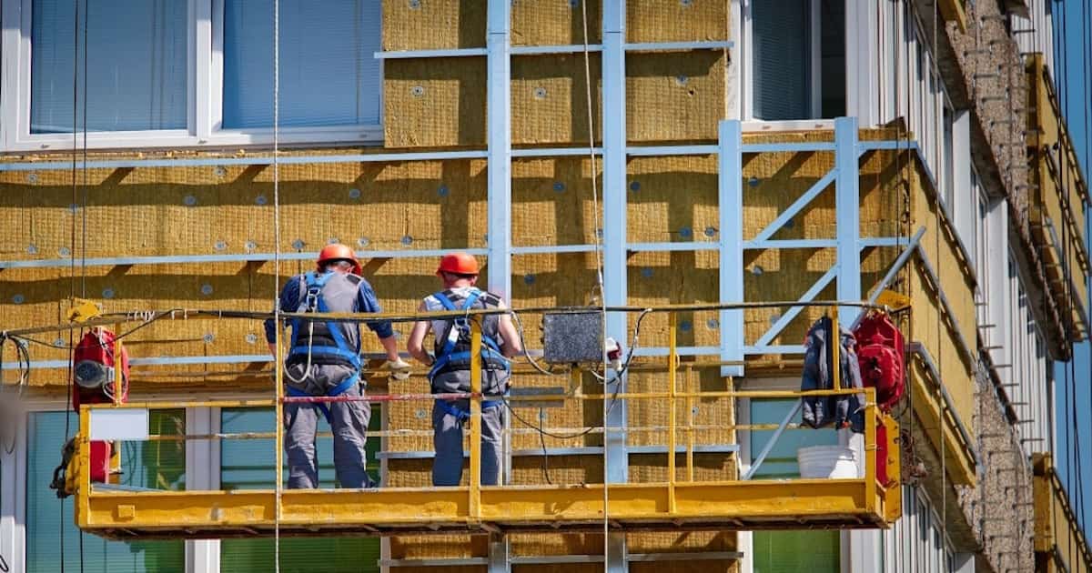 Twee gevelwerkers plaatsen gevelisolatie. Ze dragen een oranje veiligheidshelm en staan op een steiger om op hoogte te kunnen werken.