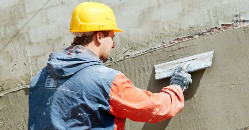 Een gevelspecialist brengt met een spaan een laag sierpleister aan over de wapeningslaag. De specialist draagt een gele veiligheidshelm. 