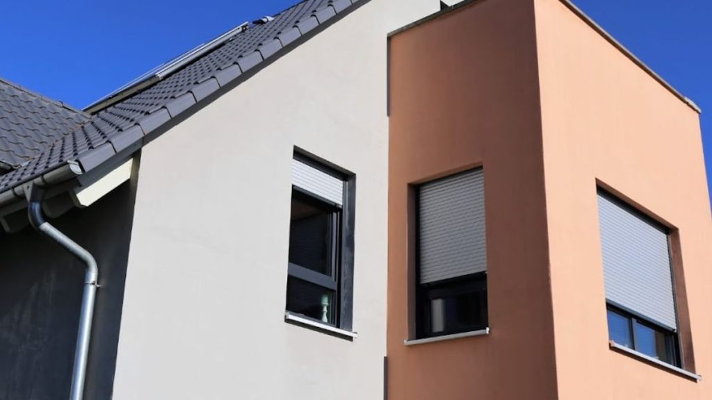 Een egale, witte gevelpleister gevel met uitstekend gedeelte dat bekleed werd met strakke, zalmroze gevelpleister. De woning heeft een donkergrijs pannendak, en drie strakke ramen.