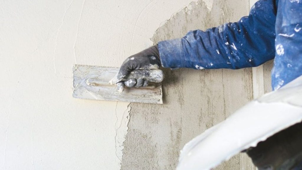 Een gevelspecialist gebruikt een pleistertroffel om dikke, witte minerale gevelpleister aan te brengen op een grijze gevel. Hij draagt een blauwe werktrui en zwarte werkhandschoenen. Zijn kleding is besmeurd met beetjes crepi.