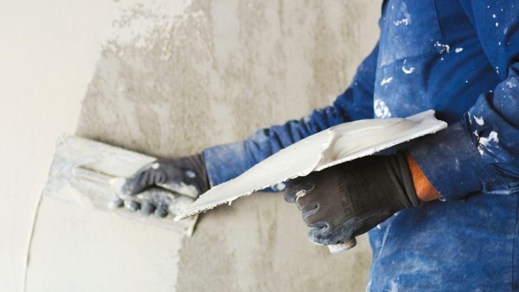 Een gevelspecialist brengt witte gevelpleister gelijkmatig aan op een grijze gevel met de hulp van een pleistertroffel. Hij draagt een blauwe trui met witte vlekjes gevelpleister, en zwarte werkhandschoenen.